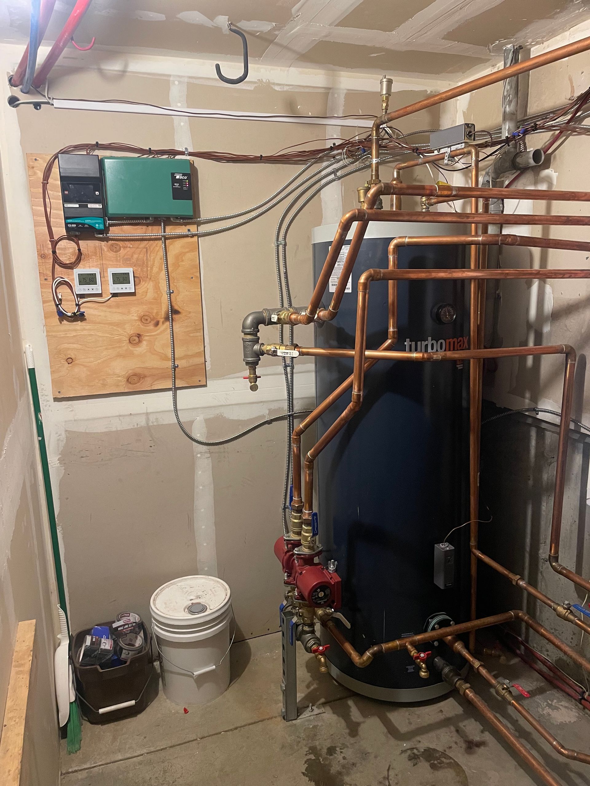 A boiler system with copper pipes, control panel, and blue tank, in an unfinished room with tools.