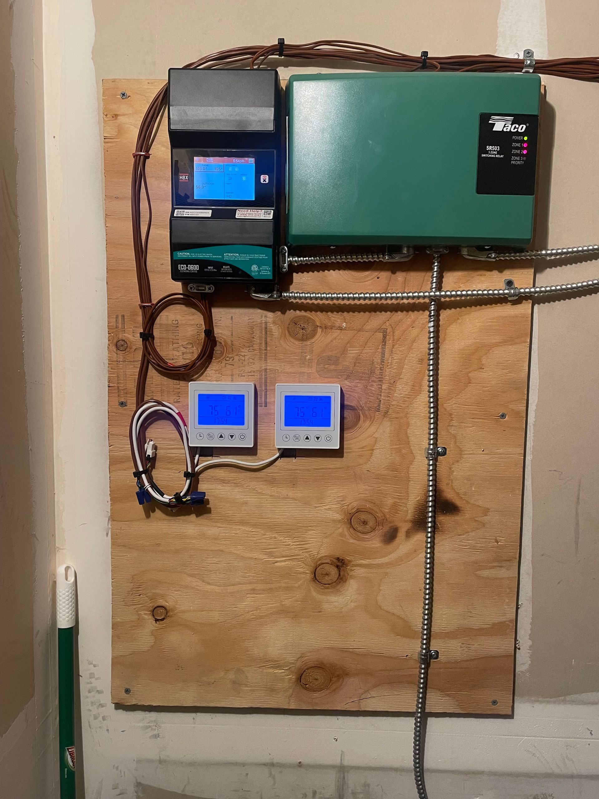 Green and black HVAC control panel with two thermostats mounted on a wooden board in a garage.