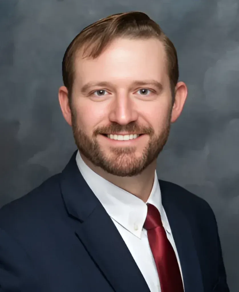 A man in a suit and tie smiles at the camera against a dark background.