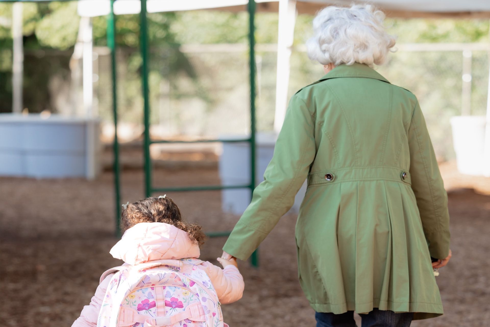 A person in a green coat walks outdoors holding the hand of a small child wearing a light pink jacket and backpack.