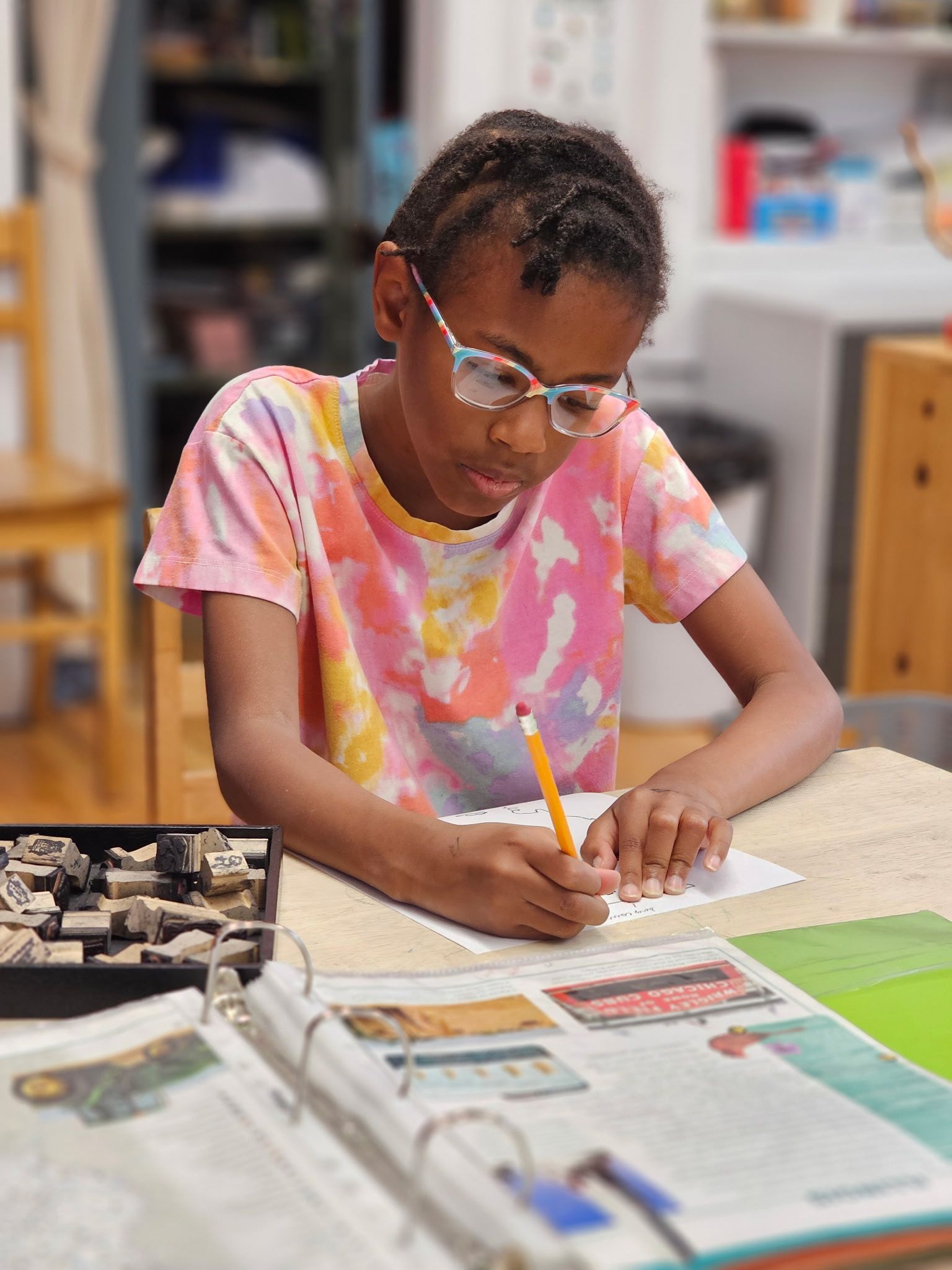 A person with glasses wears a tie-dye shirt while sitting at a desk and writing on paper with a pencil.