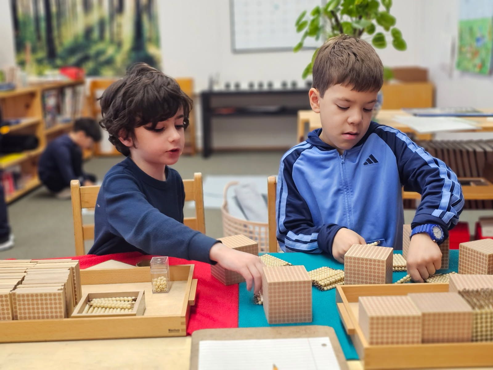 Two students in a classroom sitting at a table working with wooden base-ten blocks and math manipulatives.