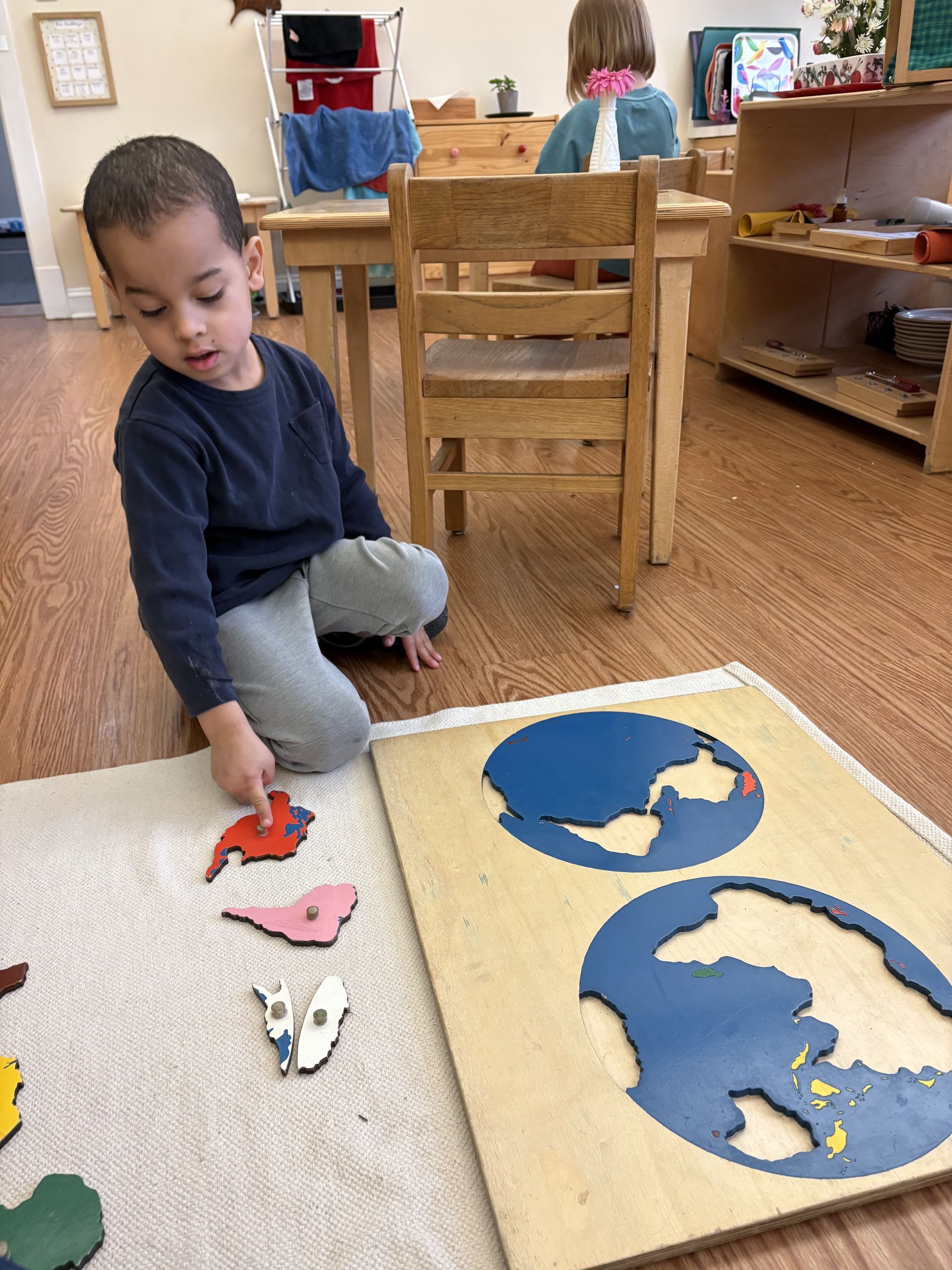 A child sits on a rug in a classroom, pointing to a continent puzzle piece while working with a world map puzzle.