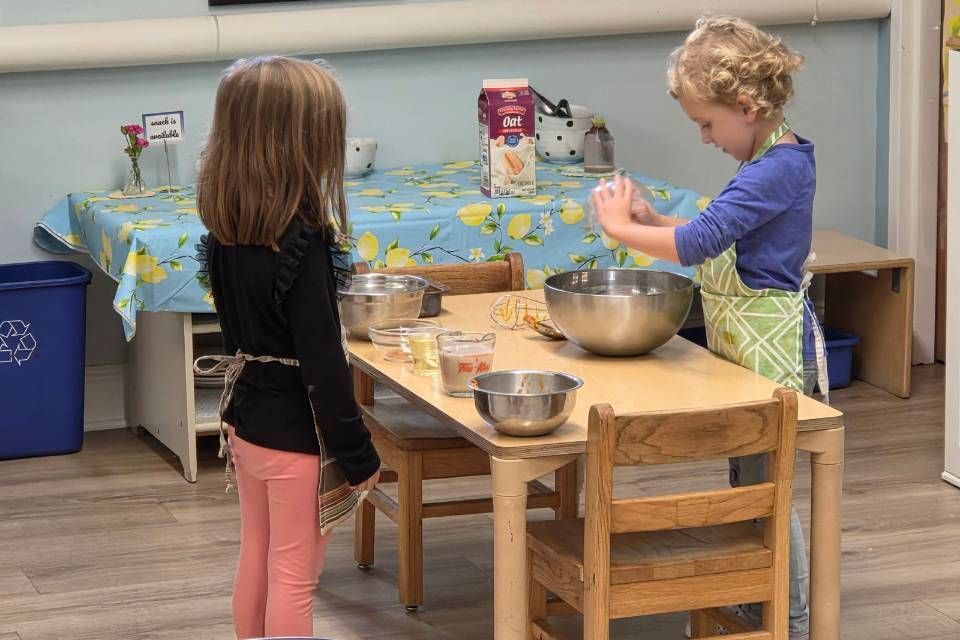Two children baking at a table. One pours ingredients into a bowl while the other watches.