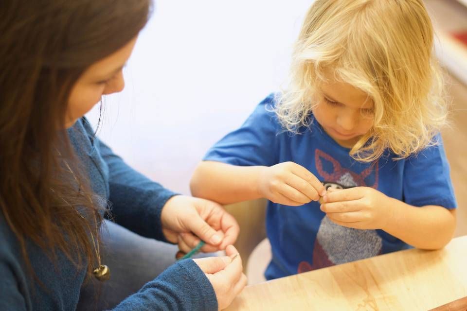 Woman and child seated at a table, stringing beads. The woman holds string, the child focuses.