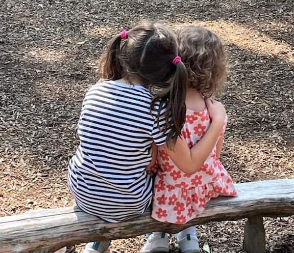 Two little girls are hugging each other while sitting on a bench.