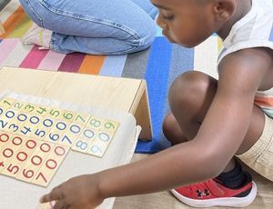 Boy playing with wooden number tiles on a rug, an adult sits nearby.
