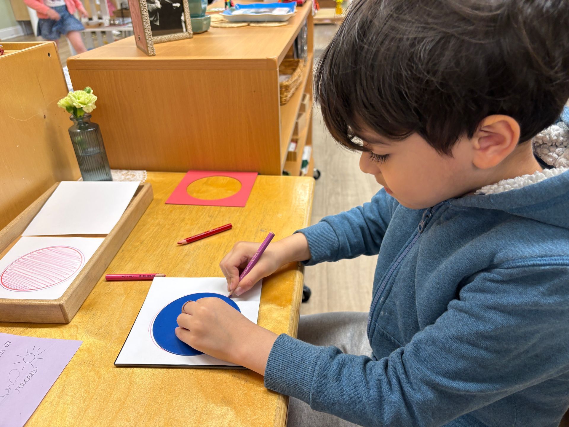 Child painting a blue circle on paper at a classroom art table
