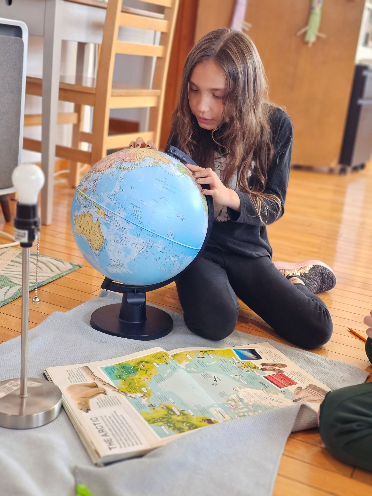Child kneeling on a rug examines a globe beside an open atlas in a classroom setting