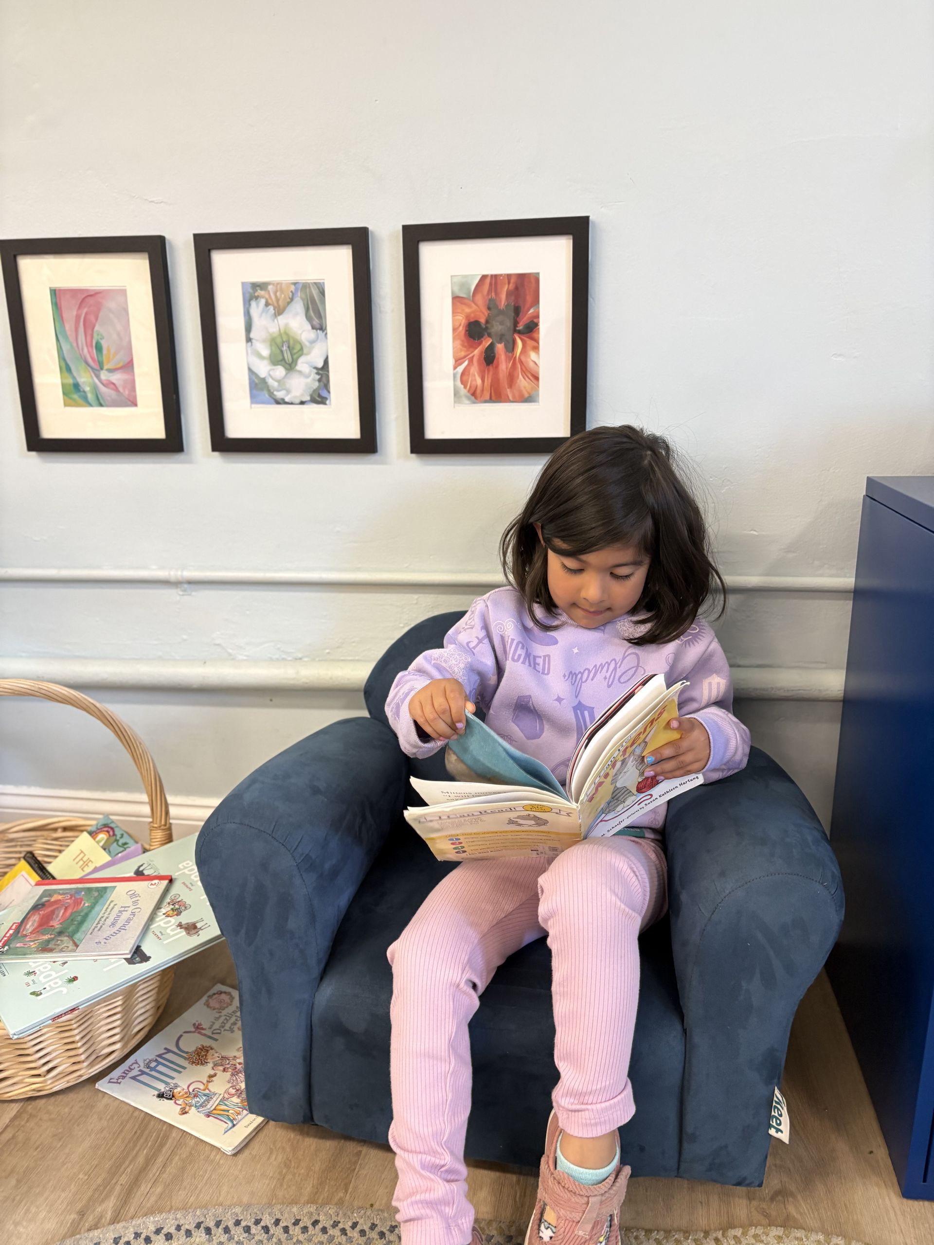 Child reading a book in a blue armchair beside framed artwork on a wall
