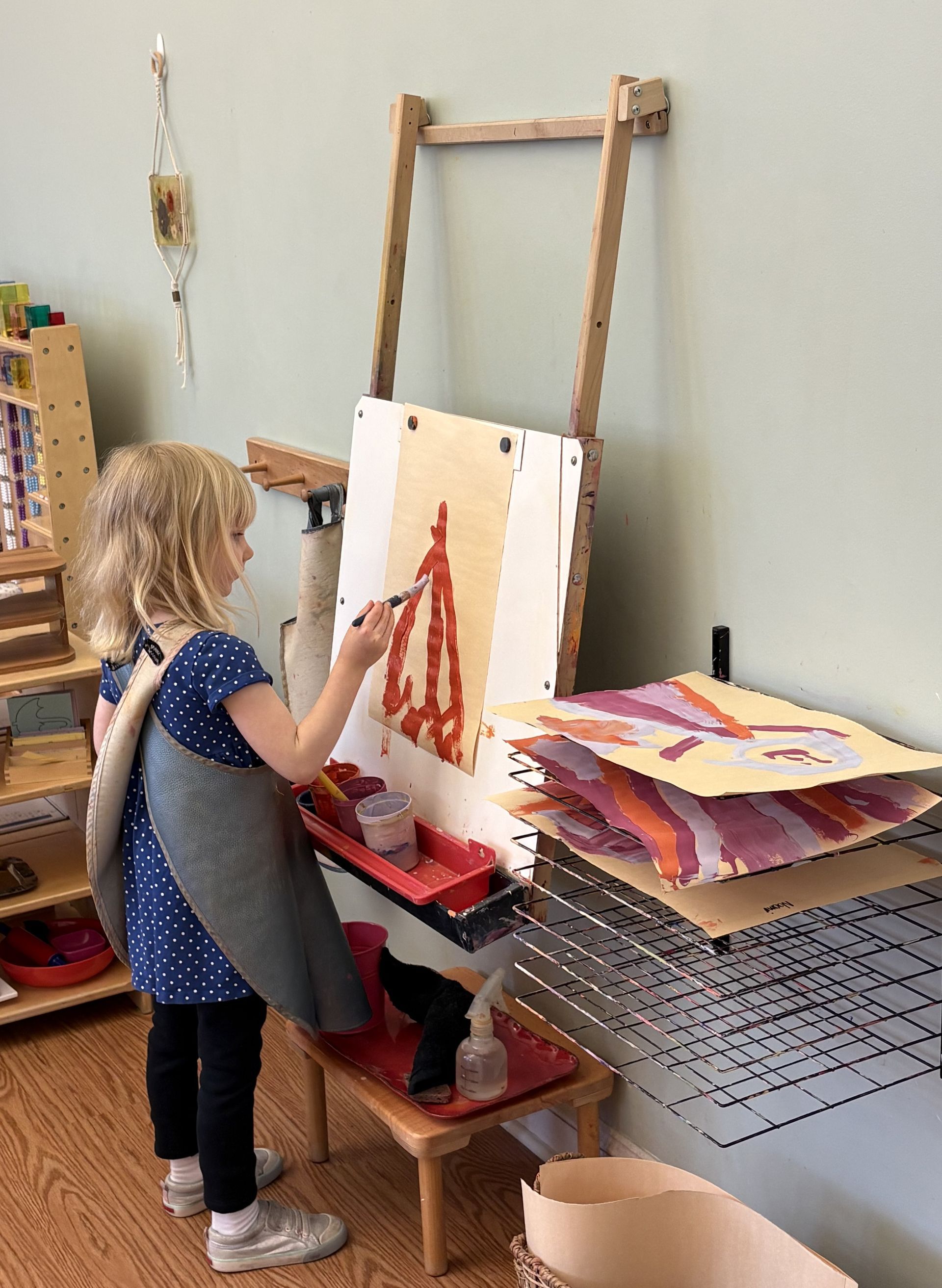 Child painting at an easel in a room, with papers and art supplies on a nearby table.