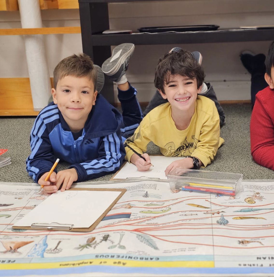 Two children lie on the floor coloring at a large Montessori timeline in a classroom.