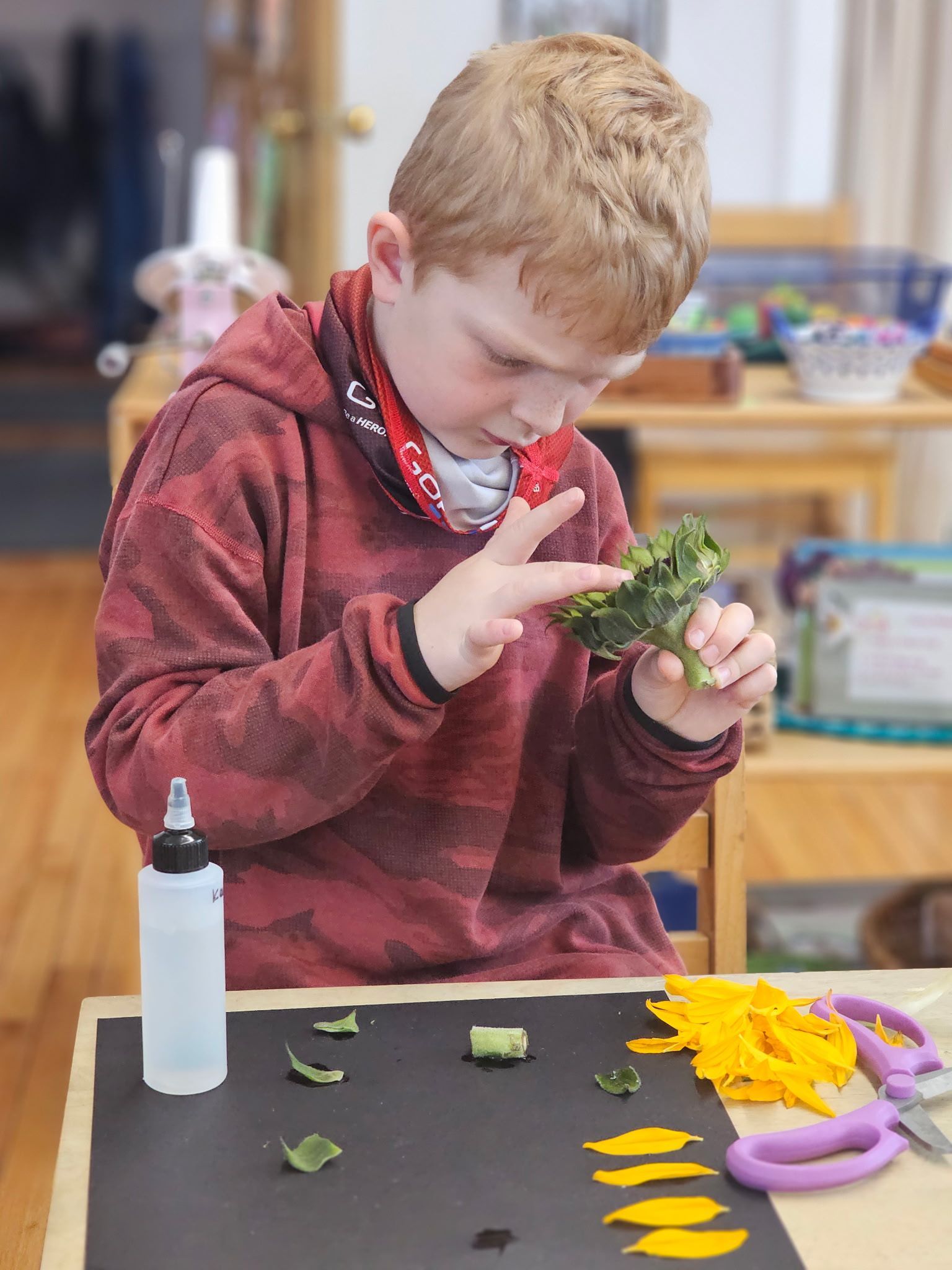 Child exploring a sunflower at a table with scissors, glue, and yellow petals