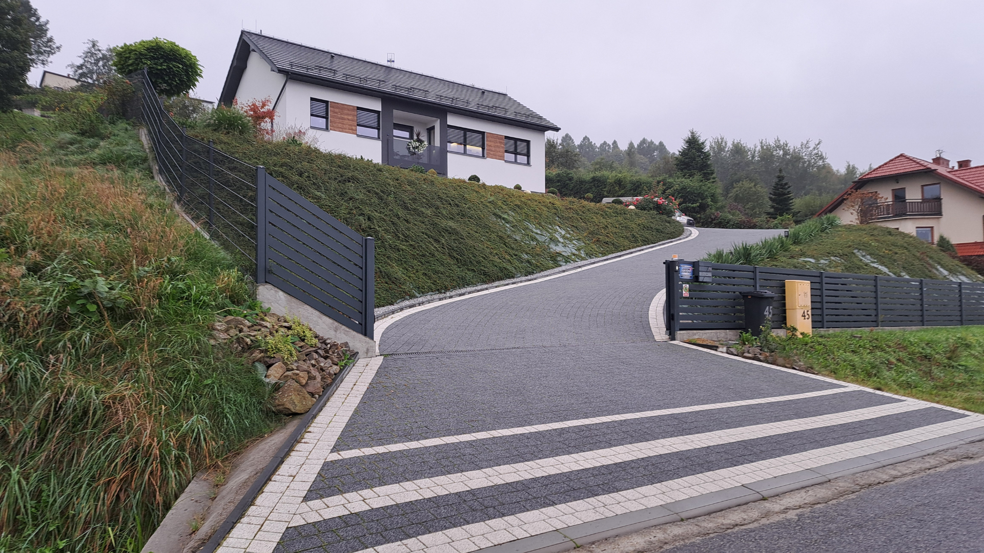 Driveway leading uphill to a white house with dark roof and trim, bordered by fences and grassy slopes.