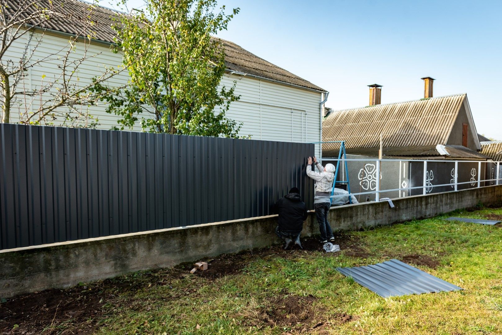 Person installing a dark gray corrugated metal fence on a concrete wall in a yard, near houses.