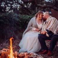 Couple sitting close by a small campfire in the woods at dusk, leaning forehead to forehead