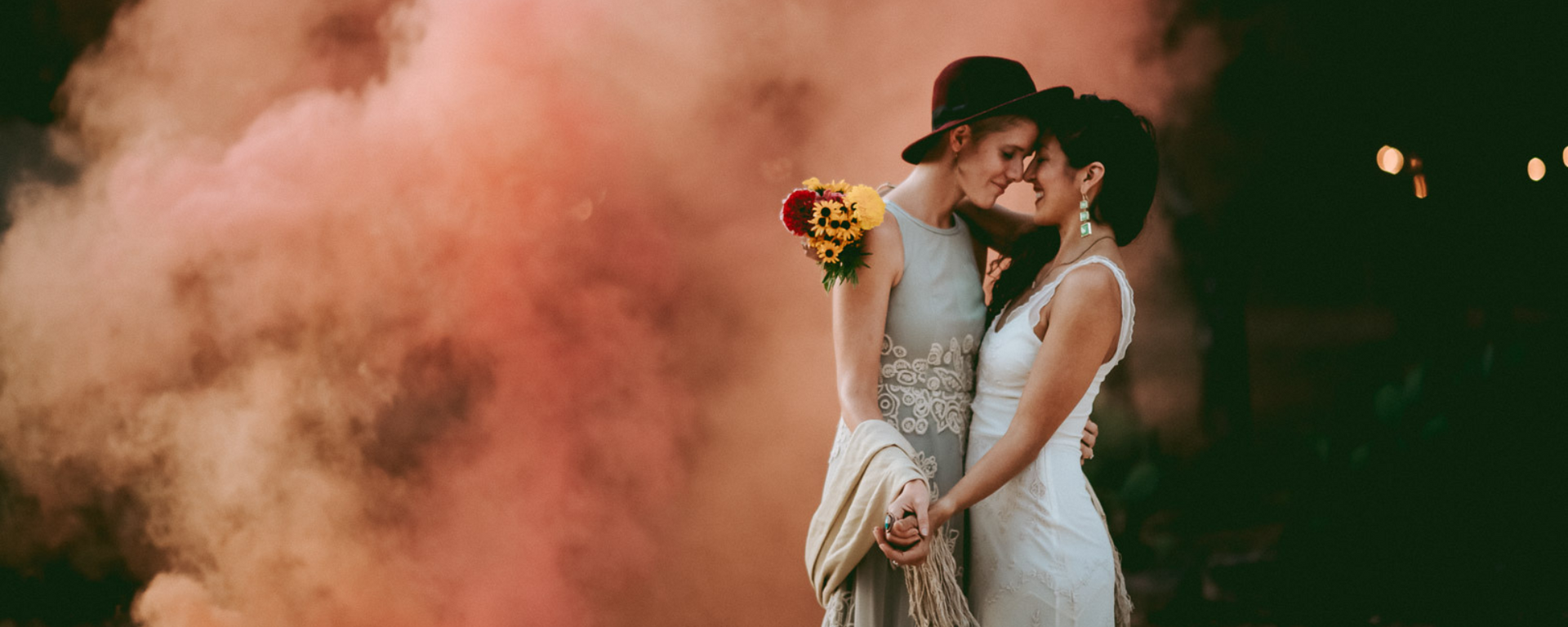 Two people in white outfits embracing amid pink smoke at night, one holding yellow flowers
