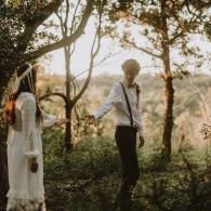 Two people holding hands in a sunlit garden, one in a white shirt and black suspenders, the other in a flowing white dress