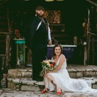 Bride in white dress seated with bouquet beside a groom in dark suit at a rustic stone wedding venue