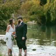 Couple in wedding attire standing in shallow water by a green riverside, smiling at each other