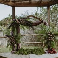 Floral wedding arch under a wooden gazebo, decorated with greenery, purple flowers, and draped fabric