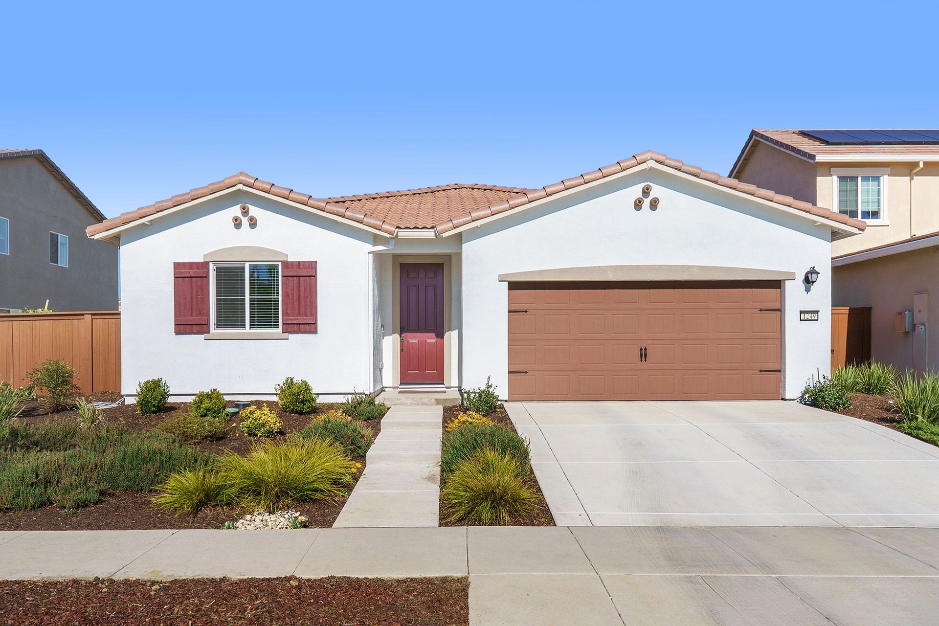 White house with brown garage door, red door, and landscaping under a clear blue sky.