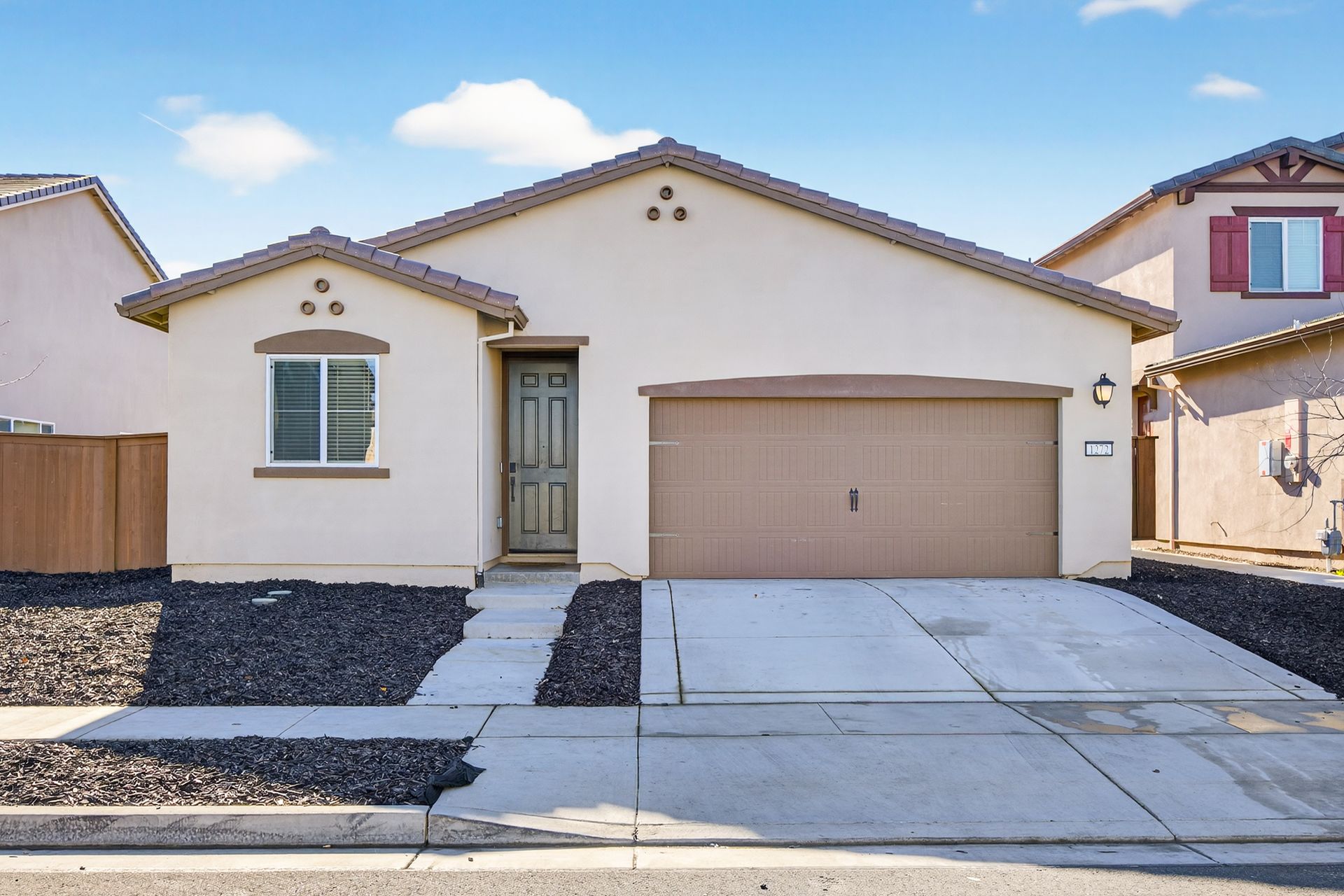 Tan stucco house with brown garage door, front porch, and gravel landscaping; blue sky.