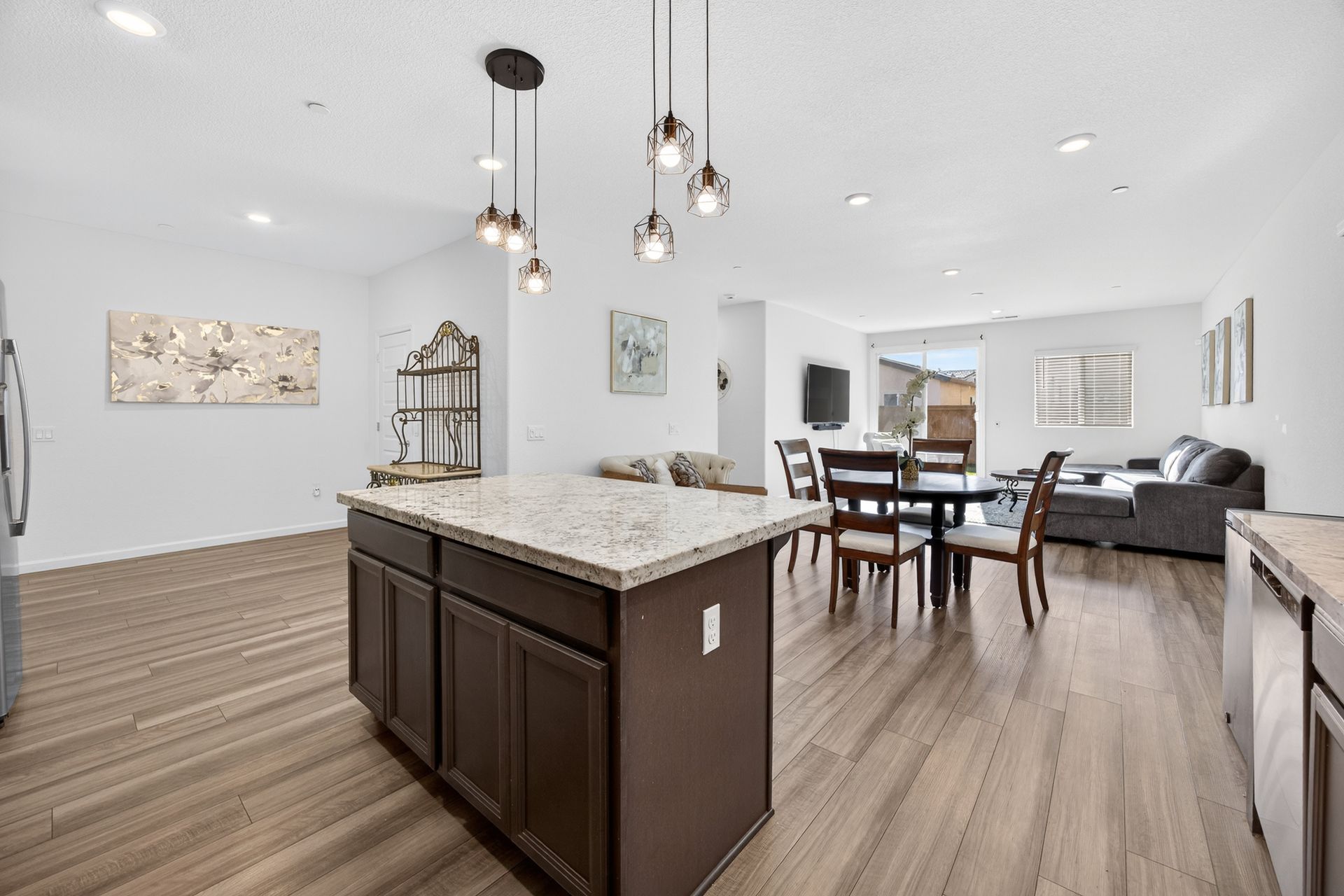 Open-plan kitchen and living area with island, pendant lights, dining table, and sofa. Wooden floors, white walls.