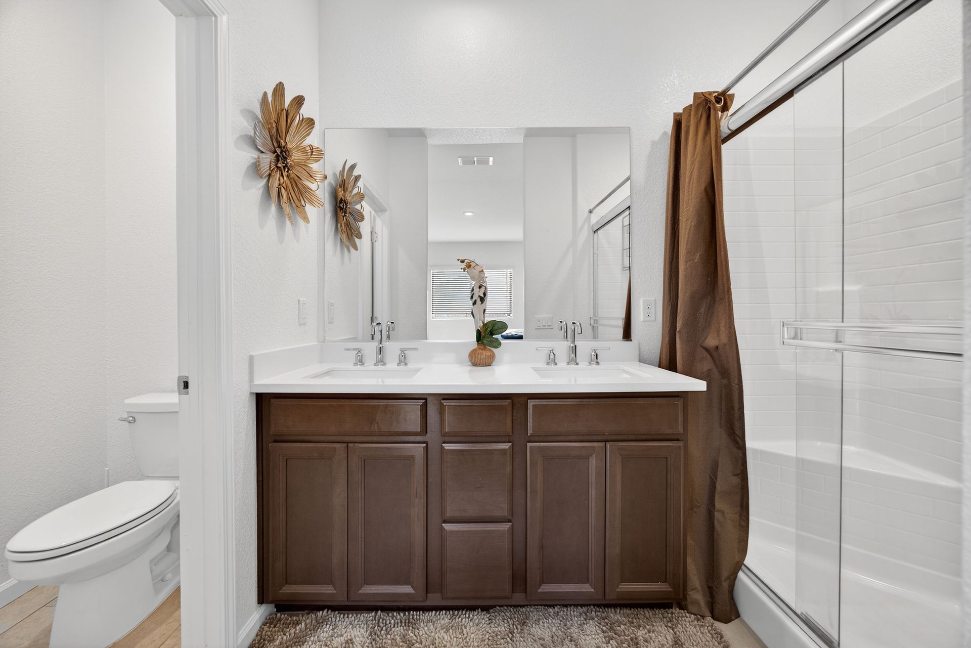 Bathroom with double sink vanity, toilet, and shower. Dark brown cabinets, white countertop, neutral walls, decorative gold wall art.