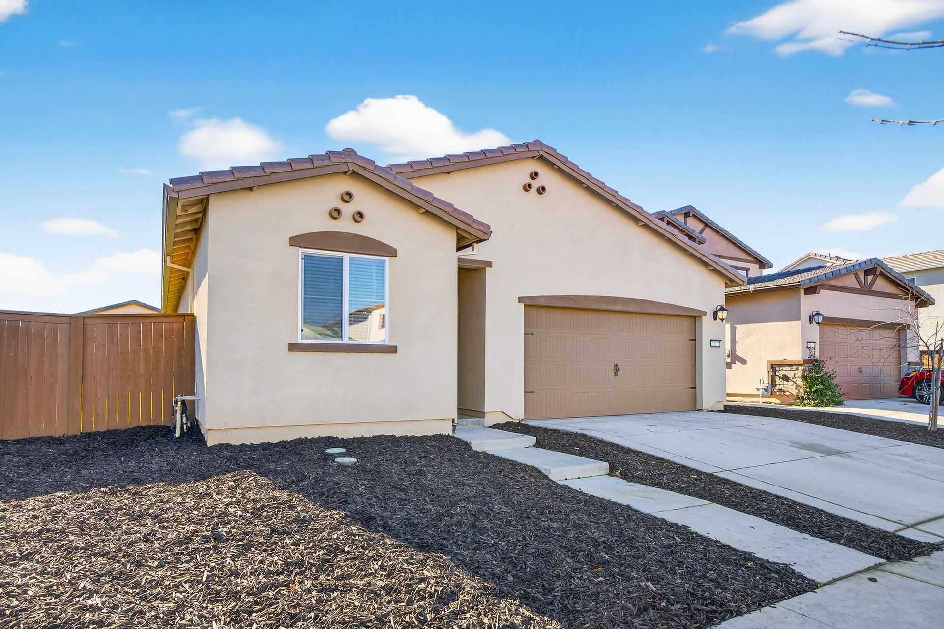 Beige stucco house with brown garage door, driveway, and landscaping on a sunny day.