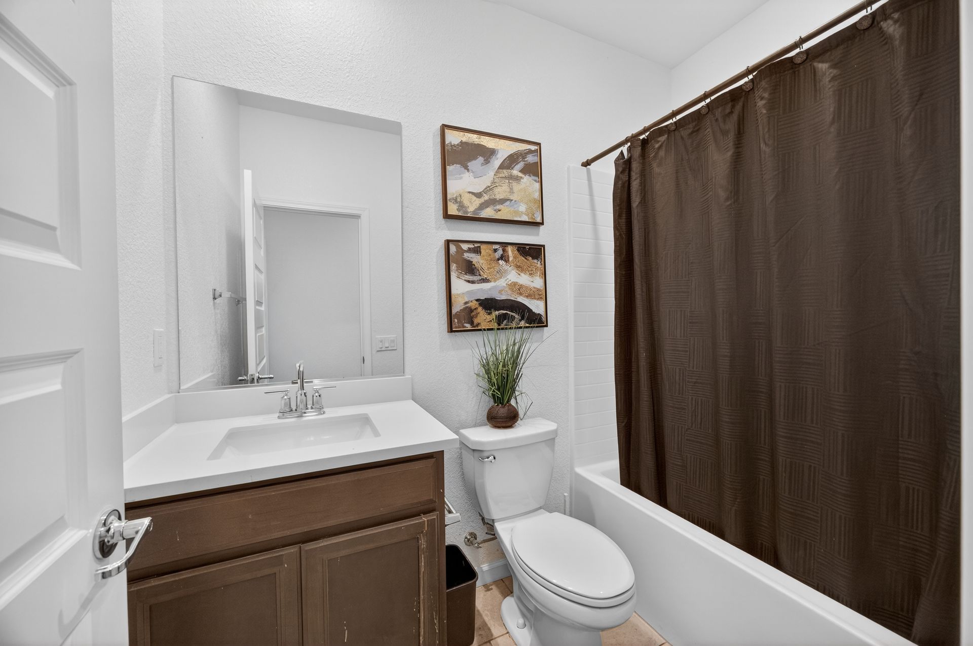 Bathroom with white walls, brown cabinet, white sink, toilet, bathtub with brown shower curtain, and framed art.