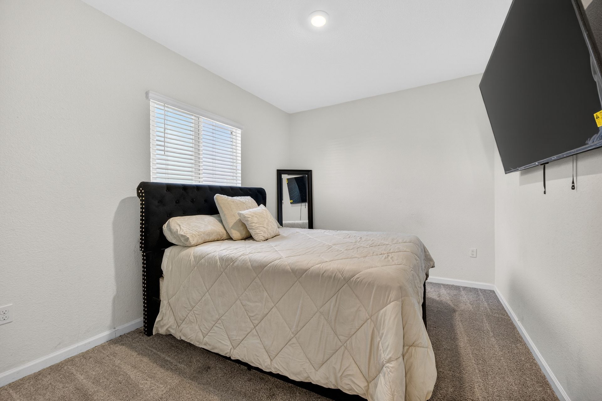 Bedroom with a bed, TV, and window. Gray carpet, white walls, and a large black TV mounted on the wall.
