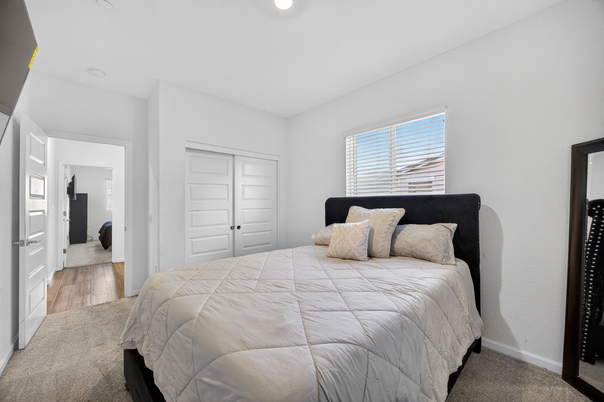 Bedroom with bed, closet doors, window, and mirror. White walls, light carpet.