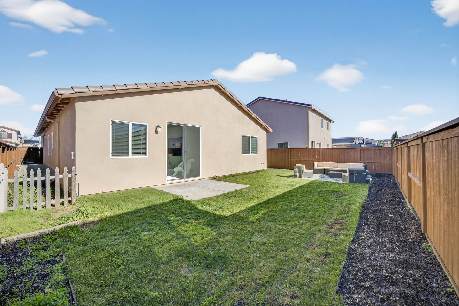 Backyard of a beige house with a lawn, patio, fence, and a clear blue sky.