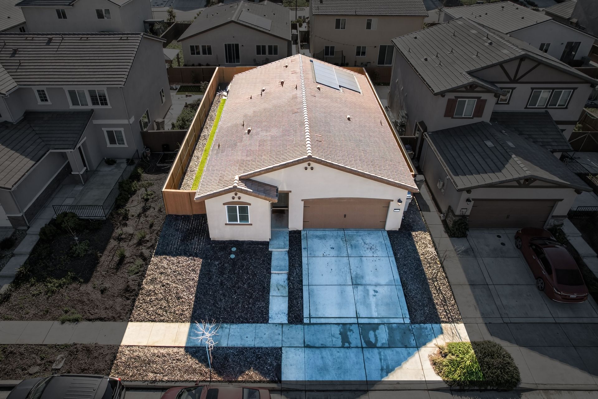 Aerial view of a single-story house with a brown roof and a driveway in a suburban neighborhood.