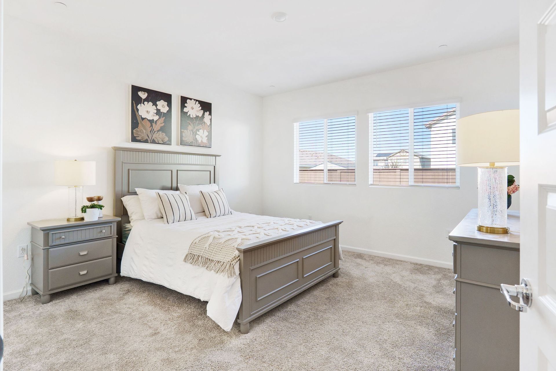 Bedroom with gray bed, nightstands, and dresser on a neutral carpet. Two windows and two floral wall art.