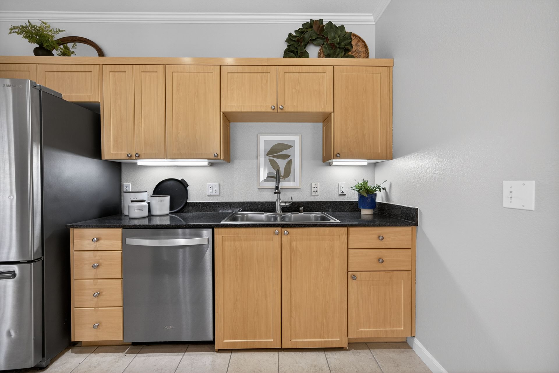 Kitchen with light wood cabinets, stainless steel appliances, dark countertop, and a small framed artwork.