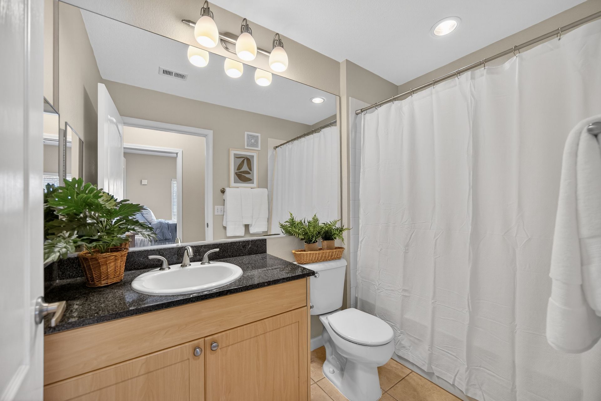 Bathroom with a sink, toilet, and shower. Light-colored cabinets and a black countertop. White shower curtain.