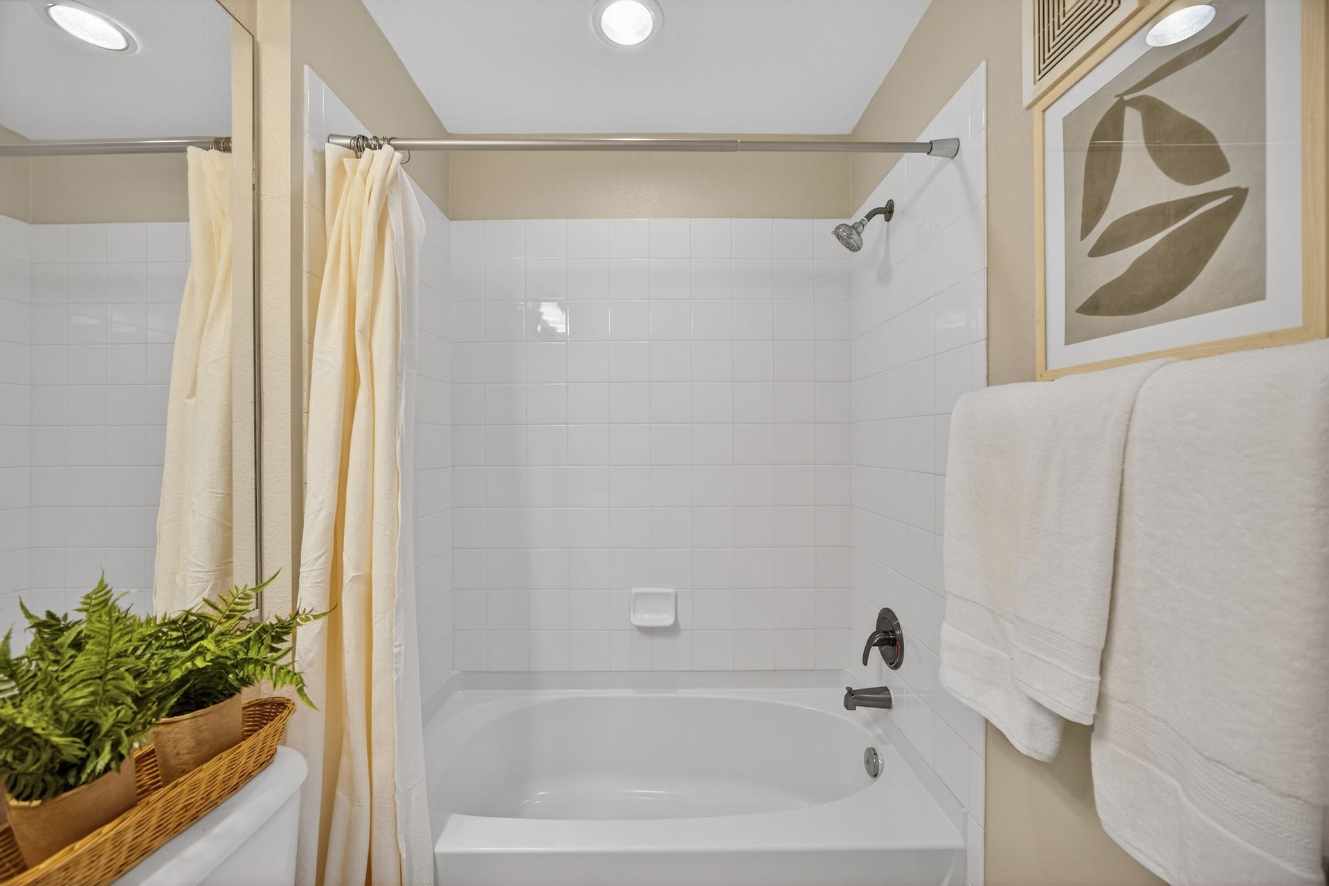 White bathroom with tub and shower, beige curtain, white towels, artwork, and potted plants.