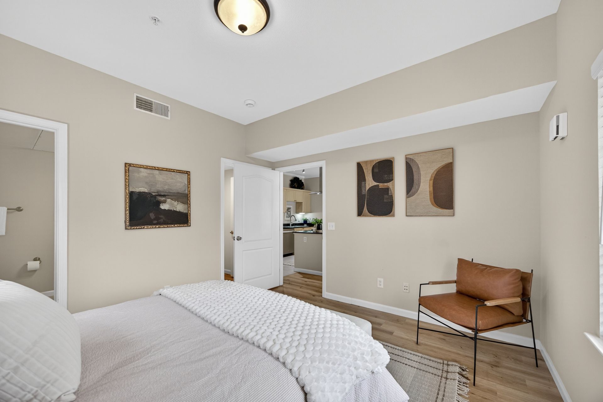 Bedroom with bed, art, chair, and open doorway to kitchen. Beige walls, wood floors.
