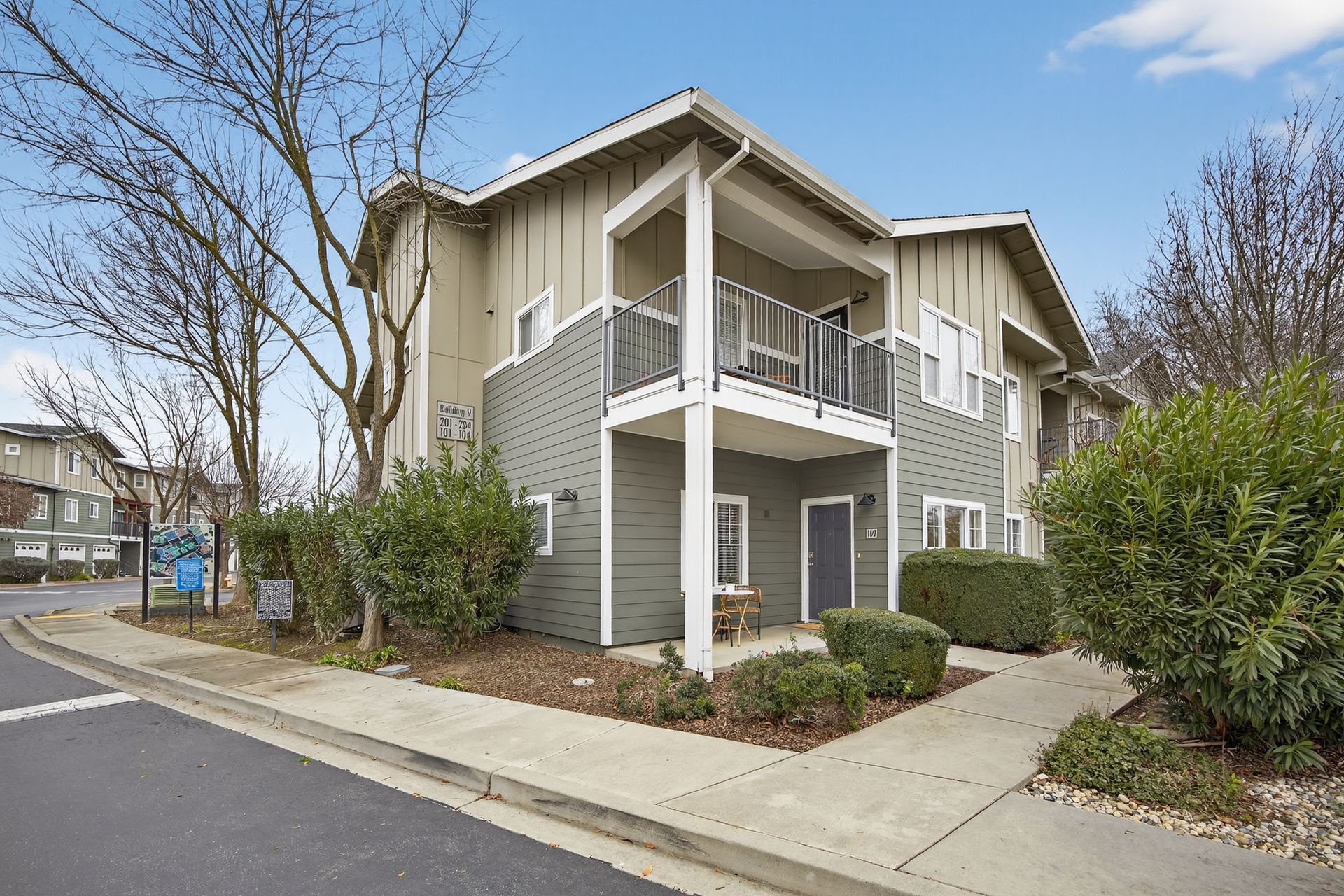 Two-story apartment building with balconies, gray siding, and a sidewalk entrance on a sunny day.
