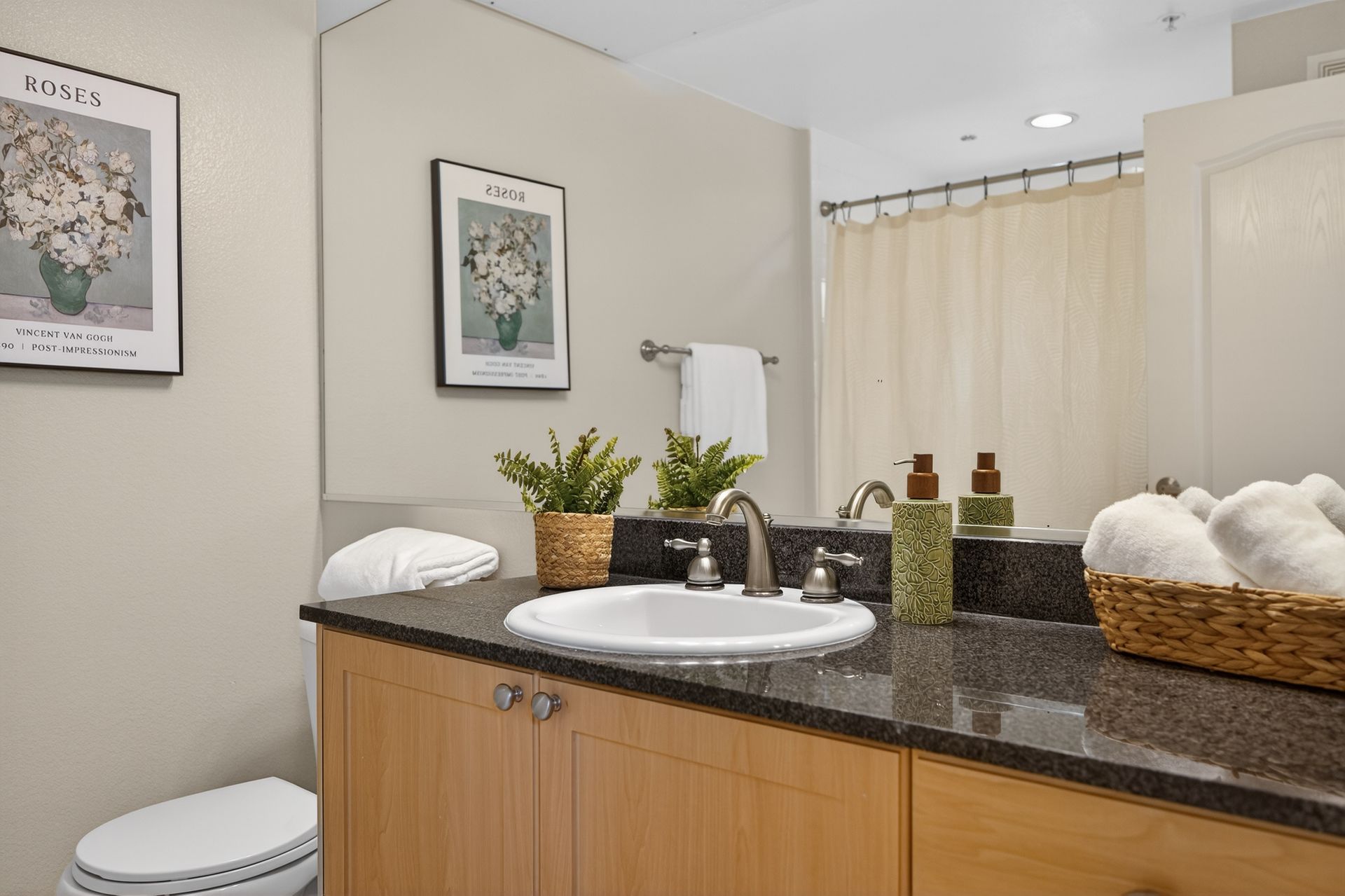 Bathroom with a sink, toilet, mirror, countertop, and framed art. Beige walls and light wood cabinets.