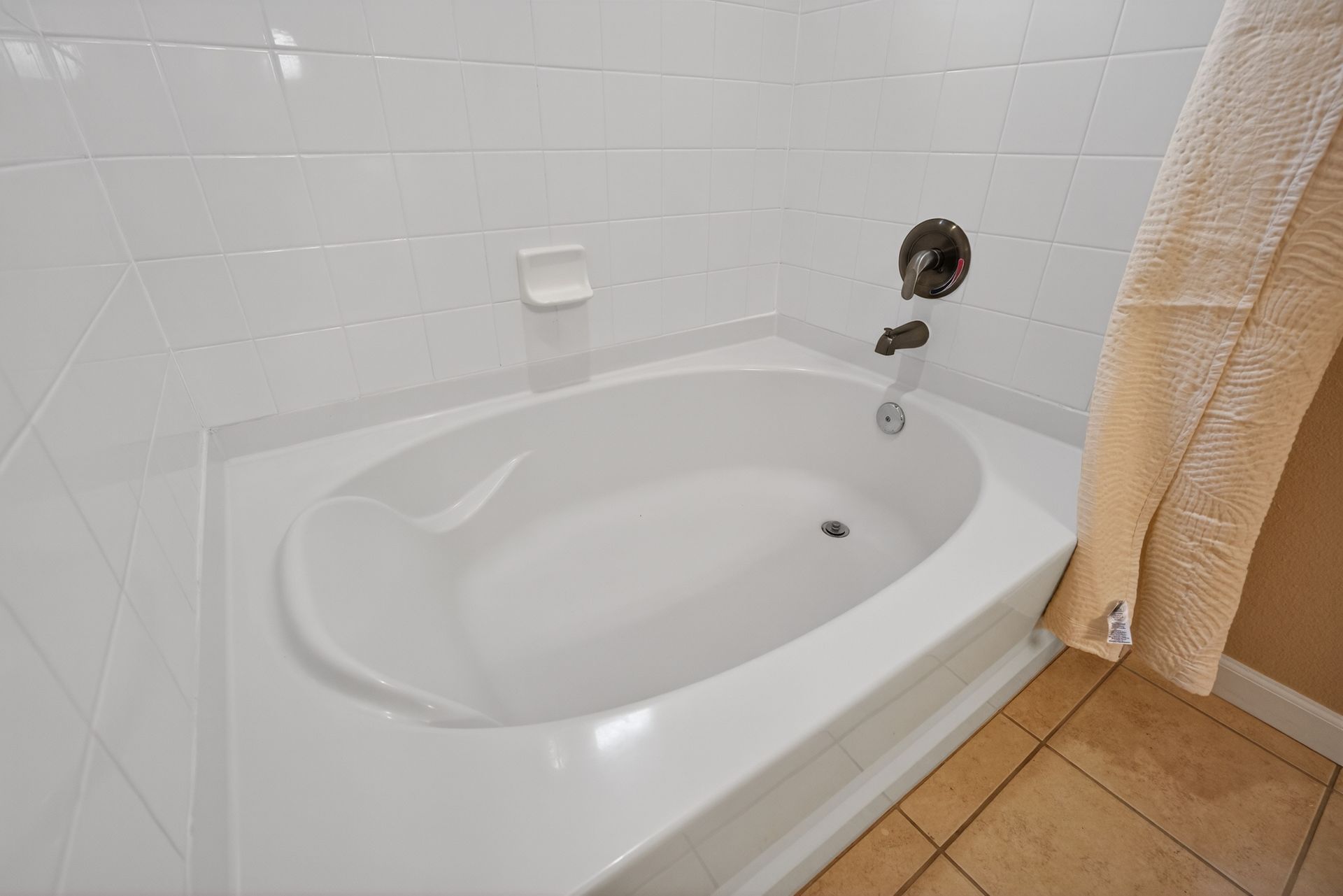 White corner bathtub in a tiled bathroom. A towel hangs from the edge.