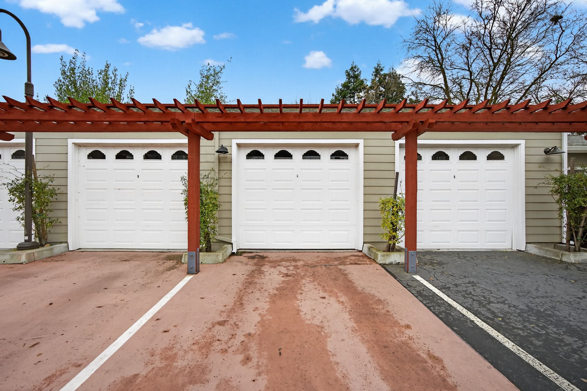 Three white garage doors with red pergola overhead, set on a red and gray driveway under blue sky.