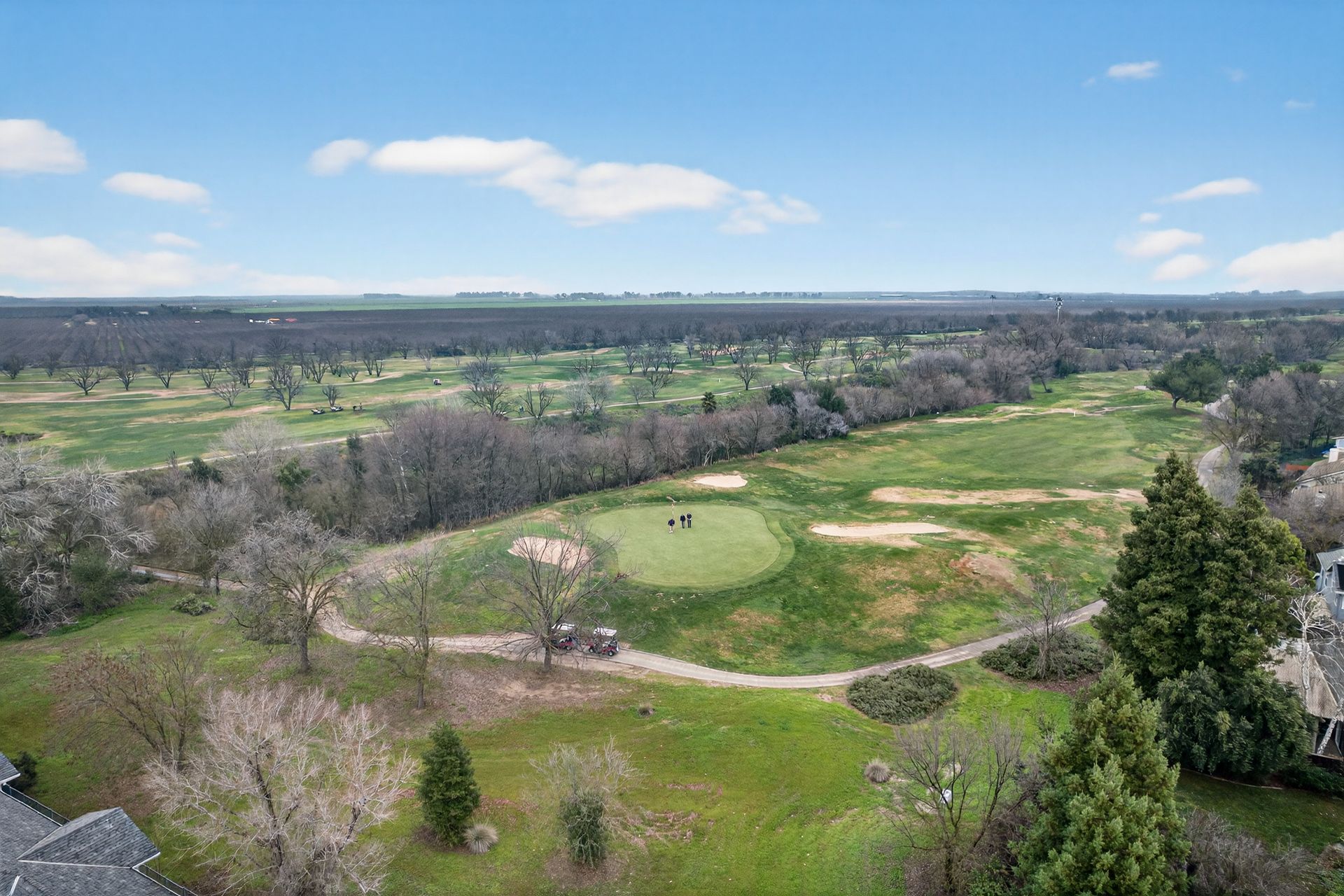 Aerial view of a green golf course with a few people playing, surrounded by trees and a cloudy sky.