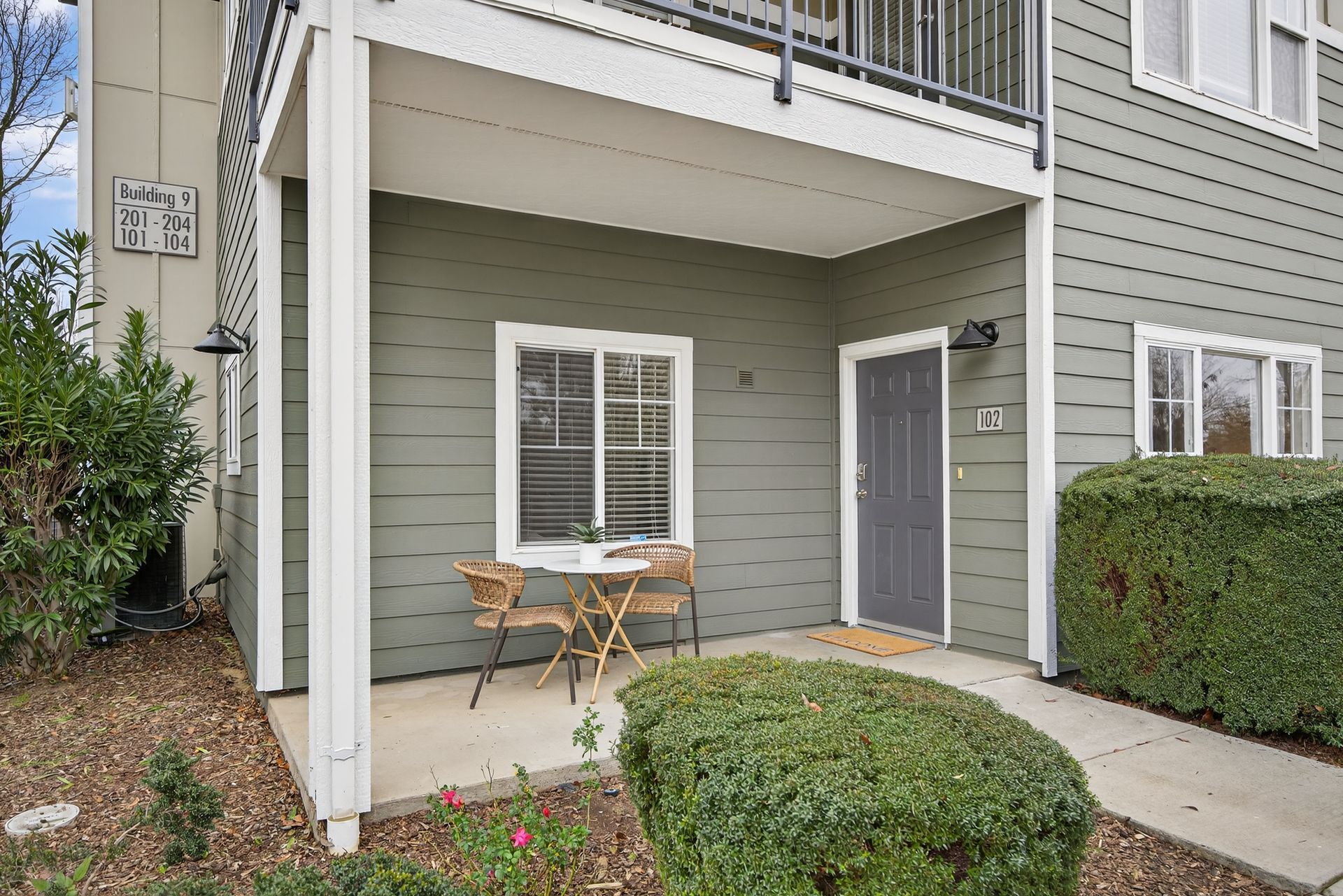Exterior view of apartment unit with small patio, table, chairs, and shrubbery.