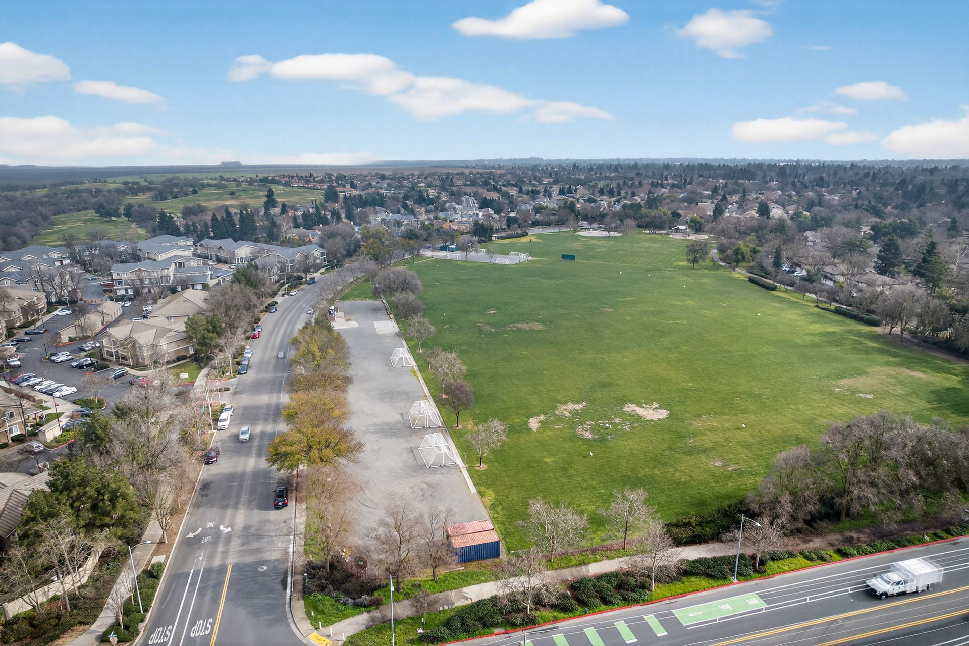 Aerial view: Road next to a grassy field, with residential buildings and trees in the distance under a blue sky.