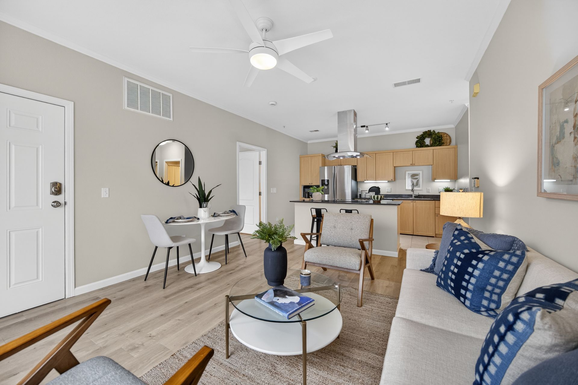Living room with neutral tones, kitchen in the background, dining area, and sofa with blue pillows.