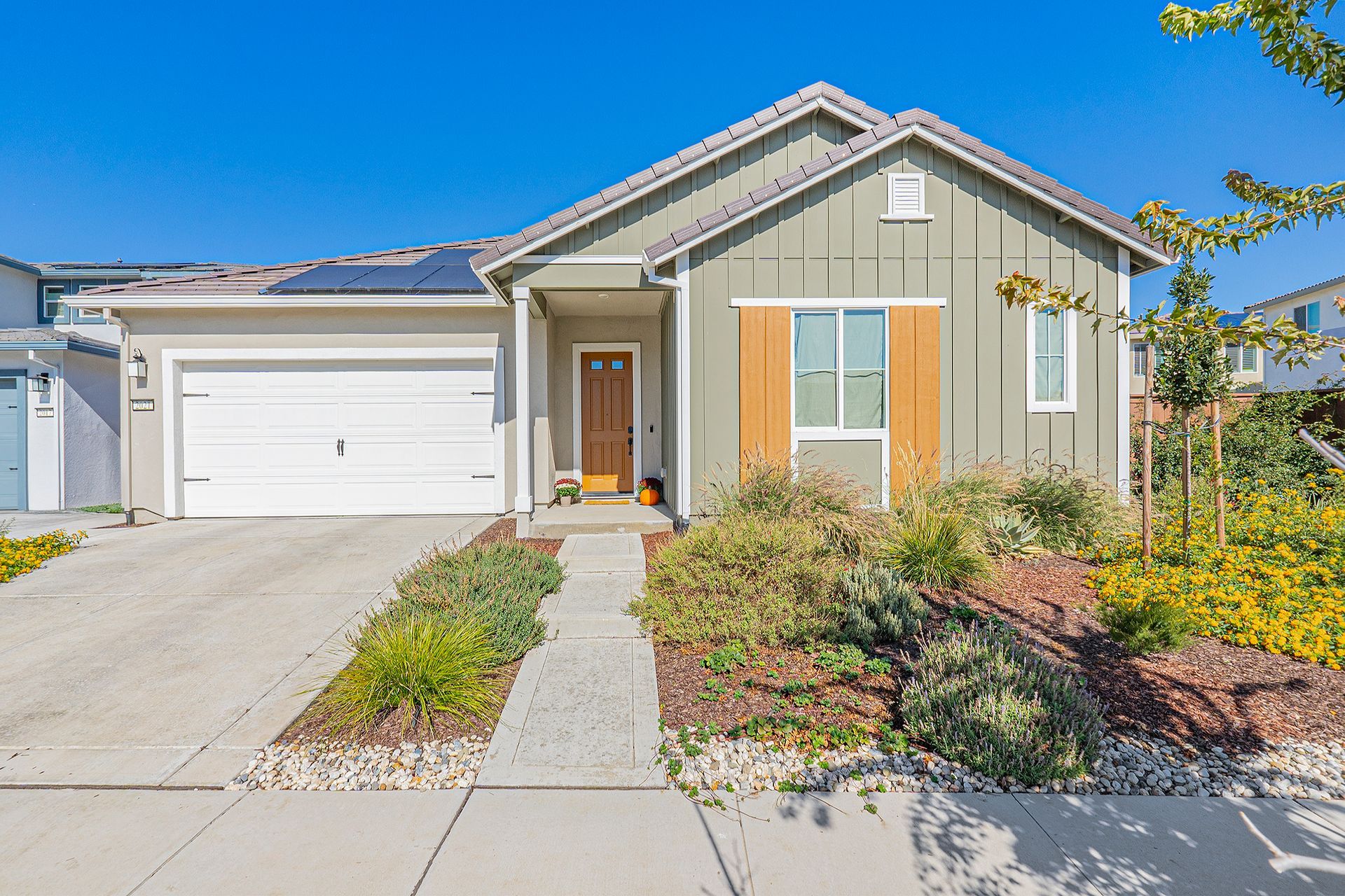 Green-sided single-story house with brown door, white garage, and pathway landscaping on a sunny day.