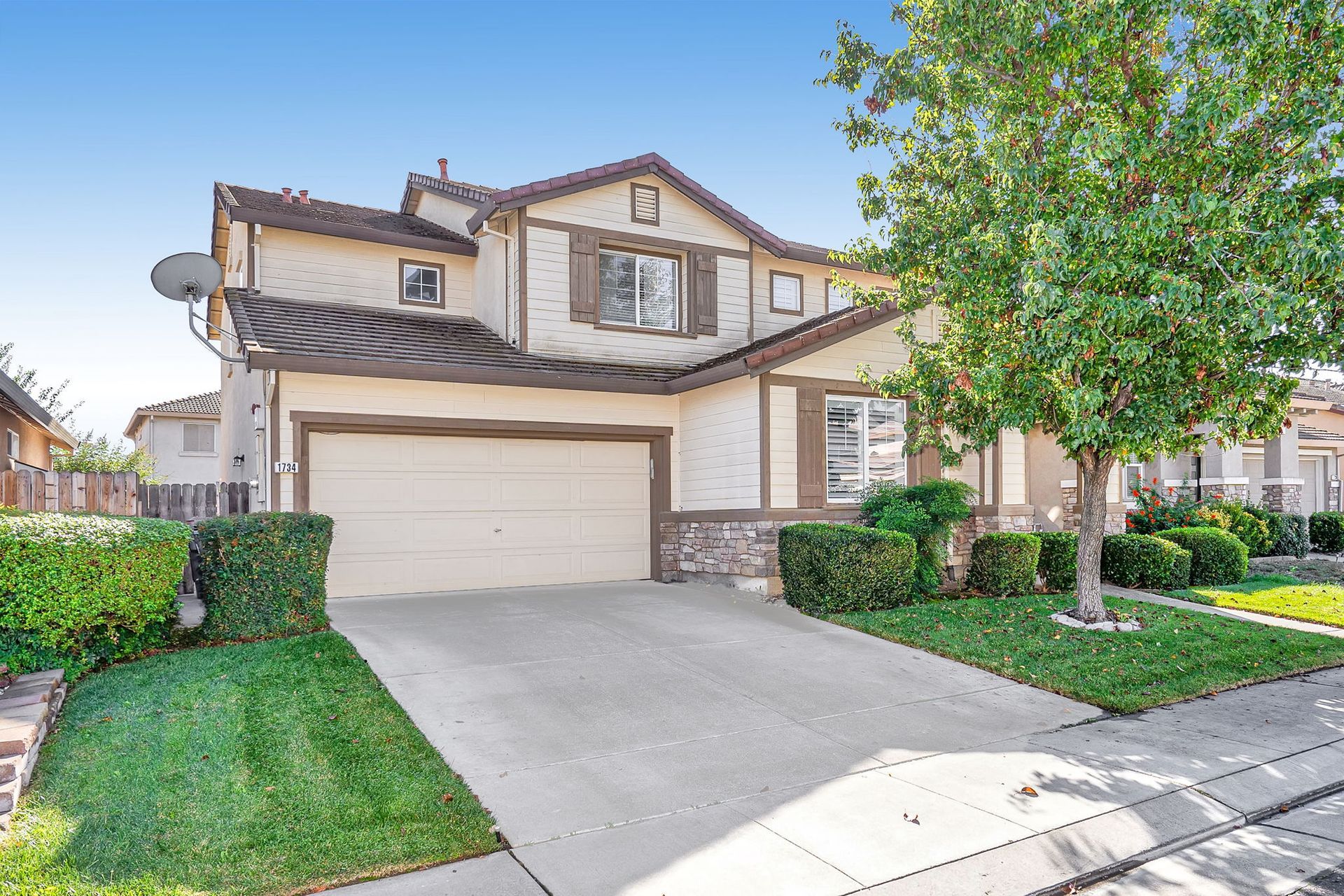 Two-story house with beige and brown exterior, driveway, and front lawn with green shrubs and trees.